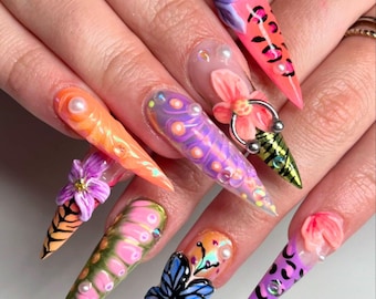 May include: Close-up of long, stiletto-shaped nails with colorful, intricate designs. The nails feature a variety of patterns, including flowers, butterflies, and animal prints. Some nails have 3D embellishments, such as pearls and flowers.