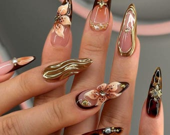 May include: Close-up of a hand with long, almond-shaped nails painted with a variety of designs. The nails feature a floral design, a gold swirl design, and a tortoise shell design. Some nails are adorned with rhinestones.