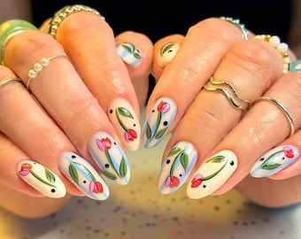 Könnte beinhalten: Ovale Nägel mit cremefarbenem Grund, verziert mit Tulpenmotiven. Jeder Nagel zeigt eine rote und rosa Tulpe mit grünen Blättern und schwarzen Punkten, vor blauen Streifen. Ein Nail-Art-Design zum Thema Frühling.