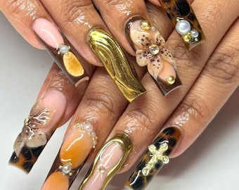 May include: Long, almond-shaped nails with various designs. Some nails feature a tortoise shell pattern, others have gold accents, and one has a 3D flower design.  A few nails have small pearl accents.