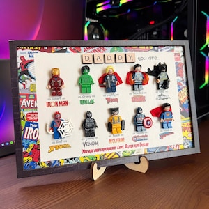 May include: A framed picture with a black frame and a white background. The picture features a collage of Lego minifigures of superheroes, including Iron Man, Hulk, Thor, Spiderman, Venom, Wolverine, Captain America, and Superman. The minifigures are arranged in rows with text below each one describing the superhero's qualities. The text reads "DADDY you are... as smart as IRON MAN, as strong as HULK, as mighty as THOR, as fast as SPIDERMAN, as funny as VENOM, as tough as WOLVERINE, as brave as CAPTAIN AMERICA, as kind as SUPERMAN. You are our superhero! Love, Olivia and Taylor."