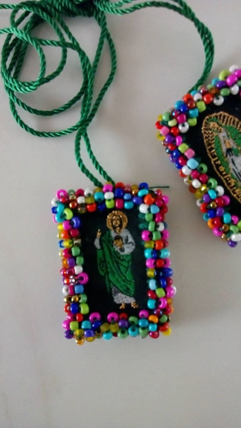 May include: Two rectangular pendants with religious imagery, framed with colorful beads. One pendant is in focus, featuring a figure in green and white on a black background. A green cord is attached. The second pendant is partially visible.