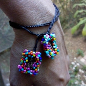 Puede incluir: Un brazalete de cordón negro con dos abalorios de cuentas de colores. Los abalorios tienen forma de rectángulo y están hechos de pequeñas cuentas multicolores.
