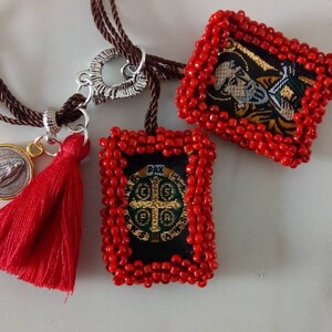 K&ouml;nnte beinhalten: Eine rote Perlenkette mit drei Anh&auml;ngern: einem silbernen Herzen, einer silbernen M&uuml;nze mit einem religi&ouml;sen Bild und einem roten Quasten. Die Kette hat auch zwei rechteckige Anh&auml;nger mit gestickten religi&ouml;sen Bildern, eines mit einem gr&uuml;nen Kreuz und das andere mit einer Figur.