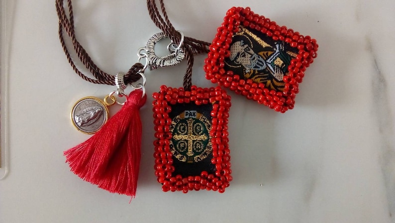 K&ouml;nnte beinhalten: Eine rote Perlenkette mit drei Anh&auml;ngern: einem silbernen Herzen, einer silbernen M&uuml;nze mit einem religi&ouml;sen Bild und einem roten Quasten. Die Kette hat auch zwei rechteckige Anh&auml;nger mit gestickten religi&ouml;sen Bildern, eines mit einem gr&uuml;nen Kreuz und das andere mit einer Figur.