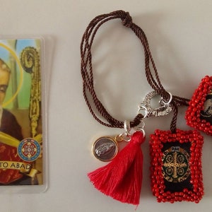 K&ouml;nnte beinhalten: Ein braunes Kordelarmband mit zwei roten Perlenanh&auml;ngern. Ein Anh&auml;nger zeigt ein kleines Bild eines Heiligen, der andere Anh&auml;nger zeigt ein Kreuz. Das Armband hat auch einen kleinen goldenen Anh&auml;nger mit einem silbernen Bild einer Feder und einem roten Quasten.