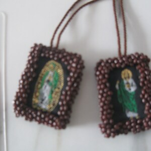 May include: Two brown beaded religious charms with images of saints. The charms are attached to brown cords.