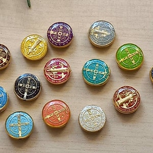 Puede incluir: Un conjunto de 13 dijes redondos de metal de colores con varios s&iacute;mbolos y dise&ntilde;os religiosos. Cada dije tiene un borde dorado y presenta una imagen diferente, incluyendo una cruz, un santo y una inscripci&oacute;n religiosa.