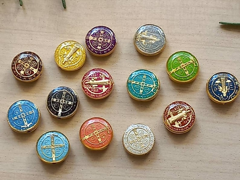 Puede incluir: Un conjunto de 13 dijes redondos de metal de colores con varios s&iacute;mbolos y dise&ntilde;os religiosos. Cada dije tiene un borde dorado y presenta una imagen diferente, incluyendo una cruz, un santo y una inscripci&oacute;n religiosa.