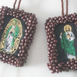 May include: Two brown beaded frames with embroidered images of religious figures. The left frame features a woman in a blue and white robe with a gold halo. The right frame features a man in a green robe with a gold halo holding a child.