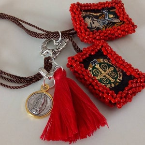 K&ouml;nnte beinhalten: Ein braunes Kordelarmband mit einem roten Quasten, einem silbernen Verschluss und zwei roten Perlenanh&auml;ngern mit gestickten religi&ouml;sen Bildern.