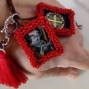 K&ouml;nnte beinhalten: Ein rotes Perlenarmband mit zwei Anh&auml;ngern. Ein Anh&auml;nger zeigt ein religi&ouml;ses Bild eines Mannes mit Bart und einem Buch. Der andere Anh&auml;nger zeigt ein religi&ouml;ses Symbol mit einem Kreuz und einem Kreis. Das Armband hat einen roten Quasten.