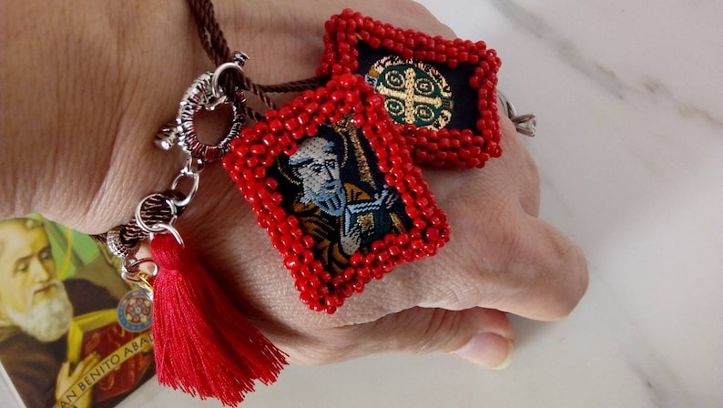 K&ouml;nnte beinhalten: Ein rotes Perlenarmband mit zwei Anh&auml;ngern. Ein Anh&auml;nger zeigt ein religi&ouml;ses Bild eines Mannes mit Bart und einem Buch. Der andere Anh&auml;nger zeigt ein religi&ouml;ses Symbol mit einem Kreuz und einem Kreis. Das Armband hat einen roten Quasten.