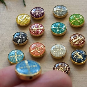 Puede incluir: Una colecci&oacute;n de 14 cuentas redondas de metal dorado con dise&ntilde;os de esmalte de colores. Cada cuenta presenta una cruz y las palabras "S.C.M.P.Q.S.V.I.B." en letras doradas. Las cuentas est&aacute;n dispuestas en c&iacute;rculo sobre una superficie de madera.
