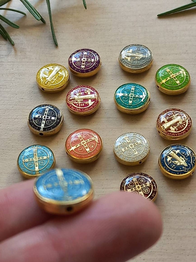 Puede incluir: Una colecci&oacute;n de 14 cuentas redondas de metal dorado con dise&ntilde;os de esmalte de colores. Cada cuenta presenta una cruz y las palabras "S.C.M.P.Q.S.V.I.B." en letras doradas. Las cuentas est&aacute;n dispuestas en c&iacute;rculo sobre una superficie de madera.