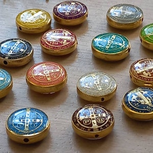 Puede incluir: Una colecci&oacute;n de 14 abalorios redondos de metal dorado con varios dise&ntilde;os de esmalte de colores. Cada abalorio presenta una cruz con detalles intrincados y fondos de colores diferentes, incluyendo azul, verde, rojo y amarillo.