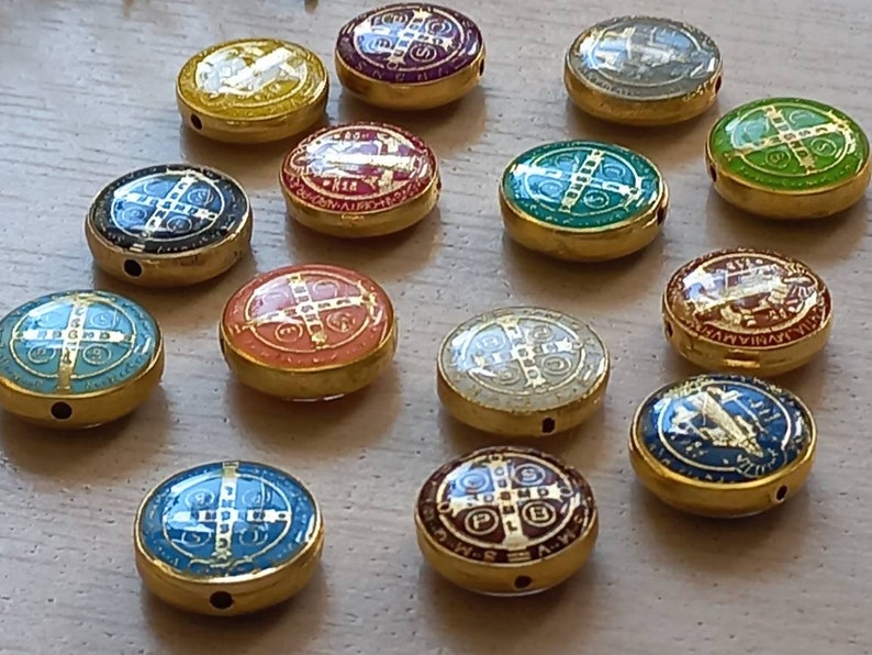 Puede incluir: Una colecci&oacute;n de 14 abalorios redondos de metal dorado con varios dise&ntilde;os de esmalte de colores. Cada abalorio presenta una cruz con detalles intrincados y fondos de colores diferentes, incluyendo azul, verde, rojo y amarillo.