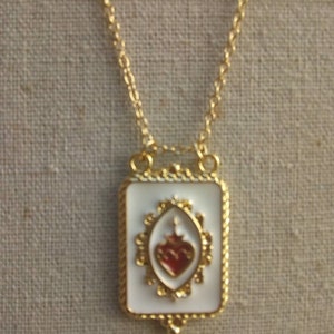 May include: Gold chain necklace with a white rectangular pendant featuring a red heart with a gold crown and ornate gold details.