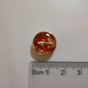 Puede incluir: Un peque&ntilde;o pin redondo rojo y dorado con un dise&ntilde;o de avi&oacute;n dorado. El pin tiene aproximadamente 1 cm de di&aacute;metro.