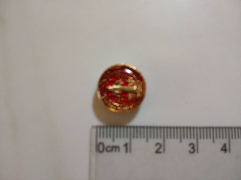 Puede incluir: Un peque&ntilde;o pin redondo rojo y dorado con un dise&ntilde;o de avi&oacute;n dorado. El pin tiene aproximadamente 1 cm de di&aacute;metro.