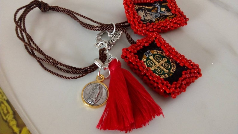 K&ouml;nnte beinhalten: Ein braunes Kordelarmband mit einem roten Quasten, einem silbernen Verschluss und zwei roten Perlenanh&auml;ngern mit gestickten religi&ouml;sen Bildern.