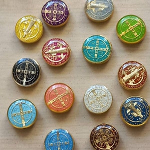Puede incluir: Una colecci&oacute;n de 14 medallas ovaladas de San Benito con ribete dorado. Las medallas son de varios colores, incluyendo rojo, azul, verde, blanco, negro y morado. Cada medalla presenta una cruz y otros s&iacute;mbolos religiosos.