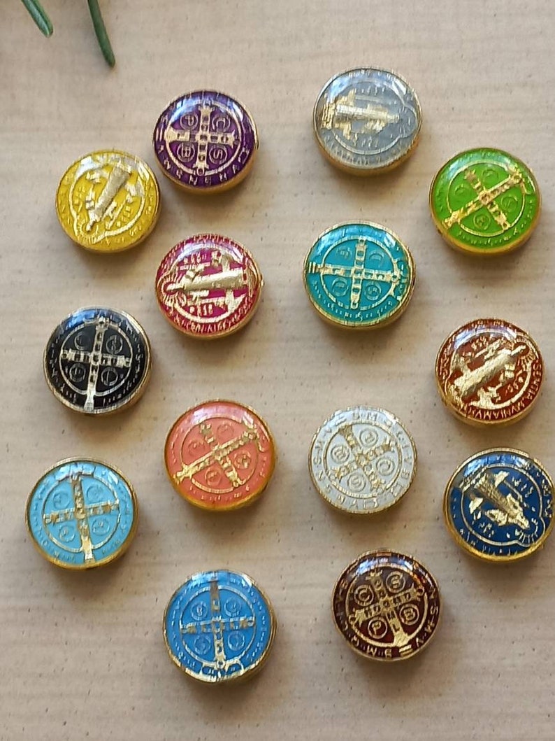 Puede incluir: Una colecci&oacute;n de 14 medallas ovaladas de San Benito con ribete dorado. Las medallas son de varios colores, incluyendo rojo, azul, verde, blanco, negro y morado. Cada medalla presenta una cruz y otros s&iacute;mbolos religiosos.