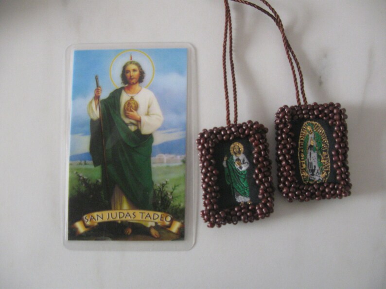 May include: Two brown beaded religious medallions with images of Saint Jude Thaddeus and Our Lady of Guadalupe. The medallions are attached to brown cords. A printed card with an image of Saint Jude Thaddeus and the text "San Judas Tadeo" is also shown.