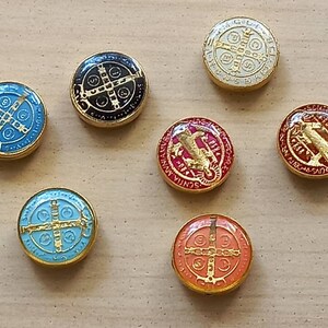 Puede incluir: Seis amuletos redondos de metal con borde dorado y centros de esmalte de diferentes colores. Cada amuleto presenta una cruz con un c&iacute;rculo alrededor y varios textos en lat&iacute;n. Los amuletos est&aacute;n sobre una superficie marr&oacute;n claro.