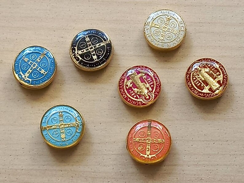 Puede incluir: Seis amuletos redondos de metal con borde dorado y centros de esmalte de diferentes colores. Cada amuleto presenta una cruz con un c&iacute;rculo alrededor y varios textos en lat&iacute;n. Los amuletos est&aacute;n sobre una superficie marr&oacute;n claro.