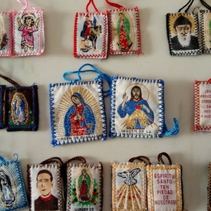 May include: A collection of 10 religious pendants with embroidered images of saints and religious figures. The pendants are made of fabric and have a variety of colors, including red, blue, green, and gold. Each pendant has a cord for hanging.