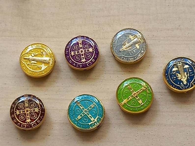 Puede incluir: Siete amuletos redondos de metal de color dorado con dise&ntilde;os de esmalte de diferentes colores. Los amuletos presentan s&iacute;mbolos religiosos y texto.