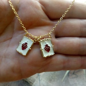 May include: A gold chain necklace with two pendants. Each pendant is a white rectangular frame with a gold border and a red heart inside.