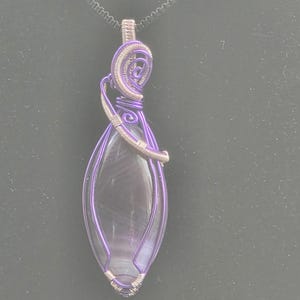 Può includere: Una collana con pendente artigianale con una pietra traslucida a forma di goccia, racchiusa in filo viola. Il lavoro del filo include spirali decorative nella parte superiore e la collana è appesa a una catena nera.