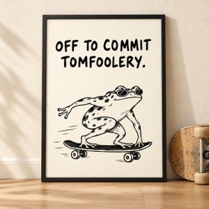 May include: A framed wall art print on a cream background featuring a black line drawing of a spotted frog wearing sunglasses and riding a skateboard. The text above the frog reads, "OFF TO COMMIT TOMFOOLERY." The frame is thin and black.