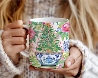 Puede incluir: Una taza de cerámica blanca con una colorida ilustración de un árbol de Navidad verde decorado con adornos rosas, colocado dentro de un cuenco con motivos azules y blancos, rodeado de flores rosas y follaje verde sobre fondo blanco.