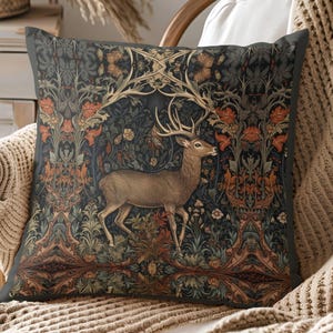May include: Decorative square pillow featuring a deer with large antlers set against a dark background with floral and foliage patterns in shades of brown, orange, and green. The pillow is placed on a chair with a neutral-colored knitted throw.