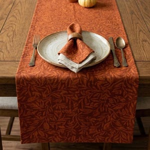 May include: A rust-colored table runner with a repeating leaf pattern, featuring a ceramic plate, a folded napkin held by a wooden ring, a fork, a knife, and a spoon, all arranged on a wooden dining table with a small pumpkin.