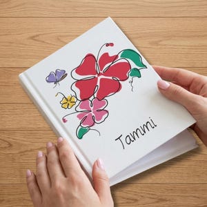 Może przedstawiać: Biały notatnik w twardej oprawie z ilustracją wykonaną ciągłą linią, przedstawiającą czerwone, różowe i żółte kwiaty z zielonymi liśćmi oraz małego fioletowego motyla. Imię Tammi jest nadrukowane czarną kursywą w prawym dolnym rogu.