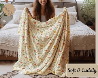 May include: A cream-colored blanket with a floral pattern featuring red and yellow flowers and green leaves. The blanket is draped over a bed. The text "Soft & Cuddly" is in a rectangular box in the lower right corner.