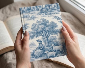 Puede incluir: Un cuaderno de tapa dura con un patrón toile de jouy en azul y blanco. El diseño muestra una escena pastoral con una persona sentada bajo un árbol grande, acompañada por dos ovejas, con una casa y follaje visibles en el fondo.