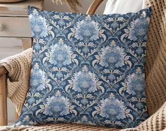 May include: Square throw pillow with a blue and white floral damask pattern. The pillow is on a wooden chair with a beige blanket. The pillow's design features a repeating pattern of stylized flowers and leaves.
