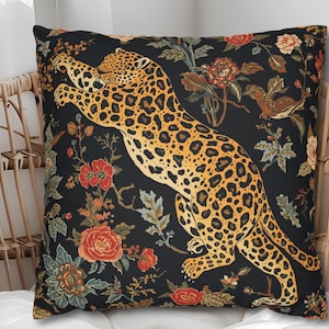 Könnte beinhalten: Quadratisches Kissen mit schwarzem Hintergrund, das einen springenden Jaguar mit gelb-braunem Fleckenmuster zeigt. Das Kissen ist mit roten, orangefarbenen und grünen Blumen- und Pflanzenmustern verziert. Es liegt auf einem Korbstuhl.