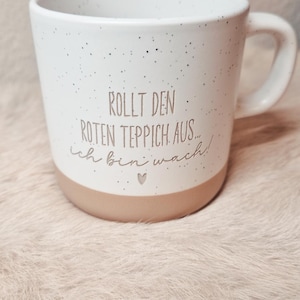 Könnte beinhalten: Weiße Keramiktasse mit braunen Sprenkeln und braunem Rand. Die Tasse hat den Text "Rollt den roten Teppich aus... ich bin wach!" in weißer Schrift.