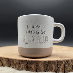 Könnte beinhalten: Ein weißer Keramikbecher mit gesprenkelter Oberfläche und einem beigefarbenen Boden. Auf der Vorderseite steht in schwarzer Schrift: Ich kaufe ein A und möchte lösen H_LTS M_UL! Der Becher hat einen geschwungenen Henkel an der rechten Seite.