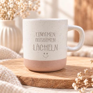 Könnte beinhalten: Ein Keramikbecher mit einem gesprenkelten weißen oberen Teil und einem einfarbigen hellrosa Boden. Der Text "EINATMEN AUSATMEN LÄCHELN" ist in Dunkelgrau auf der Vorderseite aufgedruckt, ergänzt durch ein kleines, einfaches Smiley-Symbol.