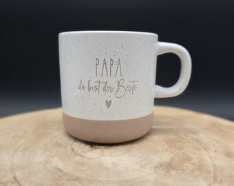 May include: White ceramic mug with a speckled finish and a tan base. The mug features the text "PAPA du bist der Beste" in a light brown color, along with a small heart symbol. The mug has a curved handle.