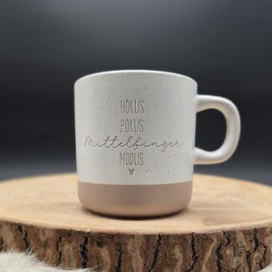 Könnte beinhalten: Eine weiße Keramik-Tasse mit gesprenkelter Oberfläche und hellbraunem Boden. Die Tasse trägt die Worte "Hokus Pokus Mittelfinger Modus" in einer verspielten Schriftart. Unter dem letzten Wort befindet sich ein kleines Herz. Die Tasse hat einen gebogenen Henkel.