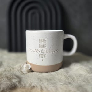 Könnte beinhalten: Weißer Keramikbecher mit gesprenkelter Oberfläche und beigefarbenem Boden. Der Becher trägt den Text "Hokus Pokus Mittelfinger Modus" in goldfarbenen Buchstaben sowie ein kleines Herzsymbol. Der Becher hat einen gebogenen Griff.