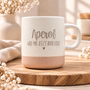 Könnte beinhalten: Ein Keramikbecher mit gesprenkeltem cremefarbenem Oberteil und einem einfarbigen hellrosa Boden. Der Text "Aperol WÄRE MIR JETZT AUCH LIEBER" ist in Dunkelgrau auf der Vorderseite aufgedruckt, ergänzt durch ein kleines Herzsymbol unter dem Text.